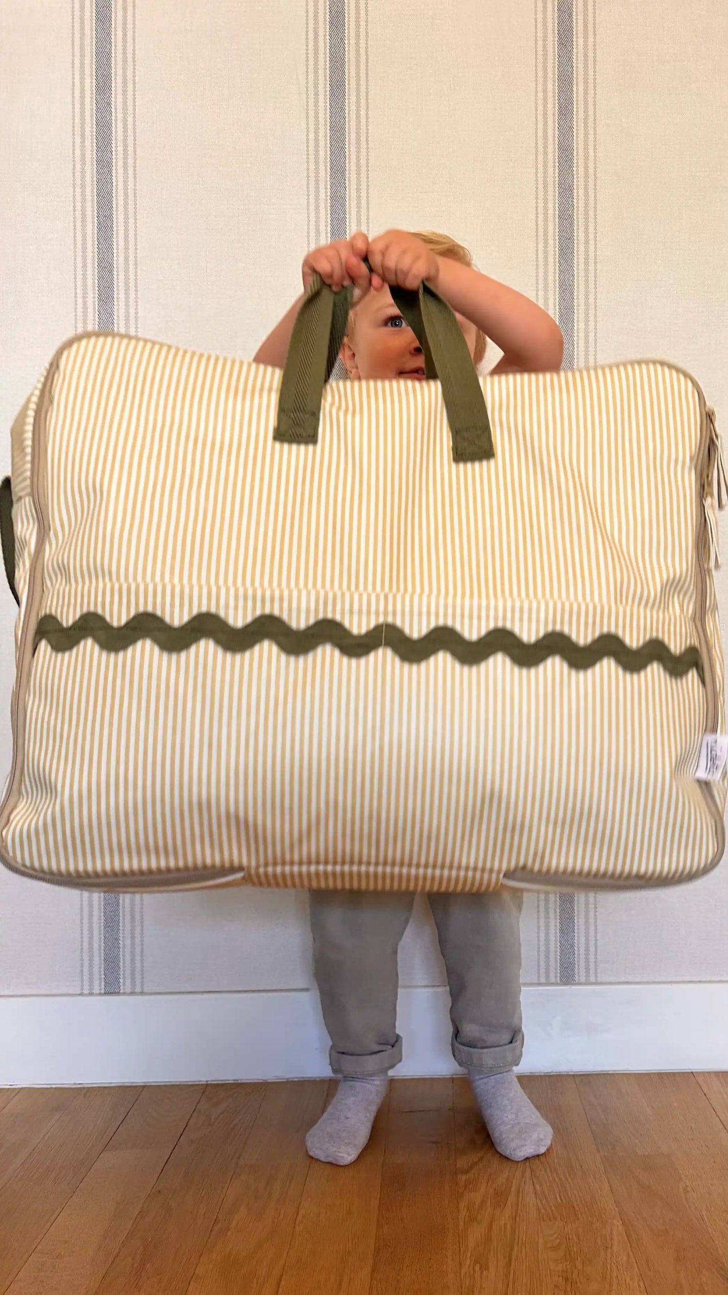 Child holding a beige and green striped maternity suitcase bag against a striped wall.