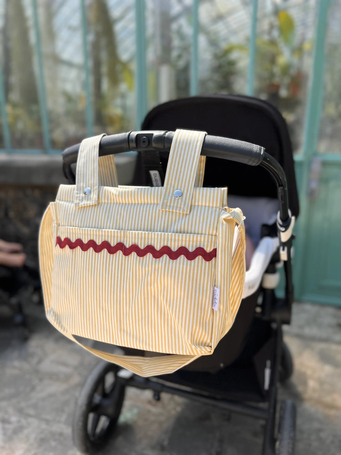 Beige stroller organizer bag with red pattern attached to a black stroller outdoors.